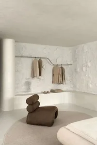 Modern interior with a white wall and concrete texture, featuring a clothing rack with beige garments and a brown sculptural 