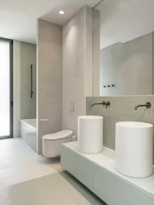 Modern minimalist bathroom featuring a white toilet, white pedestal sinks, and ceramic tiled flooring and shower wall.