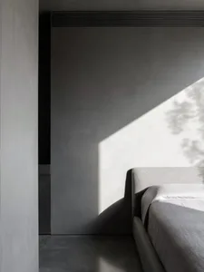 A minimalist bedroom with a concrete floor and wall, featuring a bed with a simple headboard and white bedding.