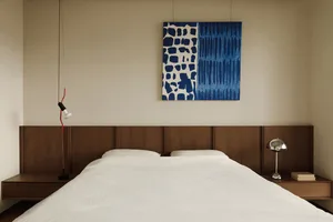 A minimalist bedroom featuring a white bed with a wooden headboard and side tables, adorned with a blue and white abstract pa