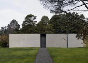 A modern, minimalist building exterior made of gray concrete with a single dark doorway surrounded by green grass and dense t