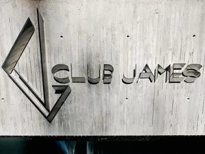 A minimalist metal sign with the text 'CLUB JAMES' on a concrete wall background.