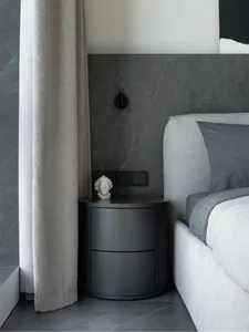 A minimalist bedroom with a concrete wall, a grey upholstered bed, a black bedside table, and a white curtain.