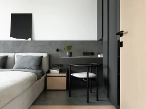 A minimalist bedroom featuring a bed with white bedding and a black headboard, accompanied by a small desk with a black chair