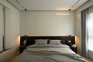 A modern bedroom with minimalist design, featuring a bed with grey bedding and black headboard, illuminated by a linear light