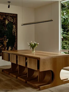 A modern dining room with a wooden table and a vase of white tulips, featuring a hanging lamp and a window with views of gree