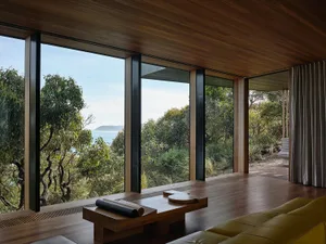 A minimalist bedroom featuring wooden flooring and a glass wall with a view of lush greenery and the ocean.