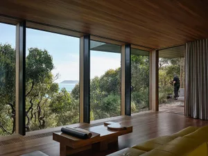 A minimalist living room with large windows overlooking a serene coastal landscape, featuring wooden furniture and a yellow s