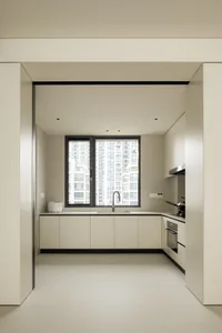 A modern minimalist kitchen with white cabinets and a large window overlooking urban buildings.
