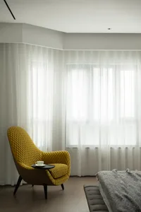 A minimalist bedroom with a yellow velvet armchair, grey bed, and sheer curtains.