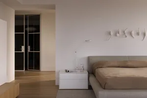 A minimalist bedroom with neutral-toned walls, furniture, and bedding, featuring a simple bed and a white lamp on a white sid