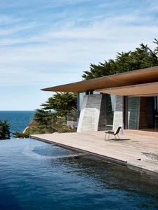 An infinity pool with a modern home exterior featuring glass walls and a wooden deck overlooking the sea.