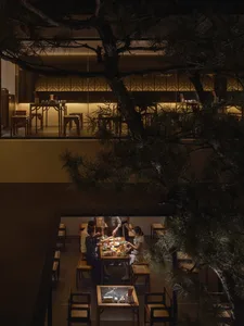A modern restaurant interior with wooden tables and chairs, illuminated by warm lighting, and featuring a tree in the backgro
