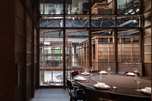 A modern restaurant dining room with a large round table and chairs, made of wood and featuring large windows.