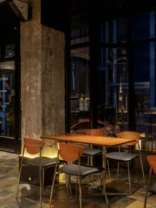 A modern restaurant interior with wooden tables and chairs, glass windows, and a stone wall.