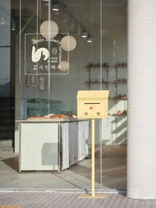 The storefront of a bakery with a light yellow mailbox in front, featuring a glass exterior and a modern design.