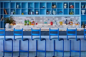 Blue chairs and metallic bar with shelves of bottles and glasses in a bar interior setting