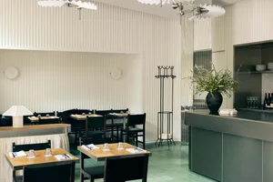 Interior of a minimalist restaurant featuring concrete walls, wooden tables, and black chairs with green plants on the bar co