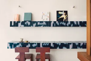 A modern coffee shop features a sleek, tiled counter with books and food items neatly arranged, and two stools for seating.