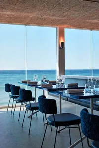 A modern restaurant interior featuring tables, chairs, and wine glasses with a glass wall overlooking the ocean.