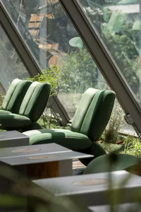 A modern cafeteria with green upholstered chairs and white tables surrounded by green plants and large windows with an outdoo