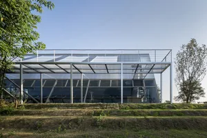 A modern glass building with a minimalist design, located outdoors with greenery in the background.