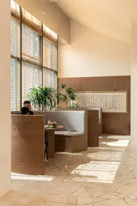 A modern restaurant interior with wooden seating, white cushioned benches, and a large window with blinds allowing natural li