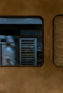 A modern restaurant kitchen with metal shelves and a window showing a baker working in the background.