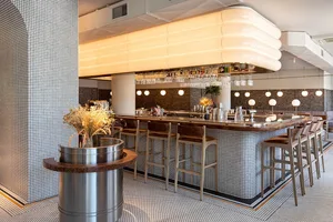 A modern restaurant interior featuring a tiled bar with sleek wooden stools and a contemporary light fixture overhead.