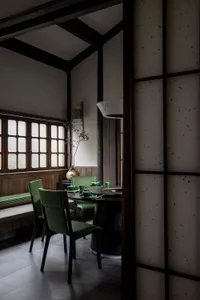 A modern restaurant interior featuring a round wooden table surrounded by green upholstered chairs, set against a backdrop of