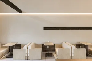 A modern minimalist restaurant featuring beige fabric seating and dark wooden accents.