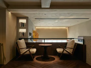 A modern reception area featuring concrete walls and floor with a round wooden table and two cushioned chairs.