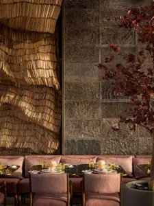 A modern restaurant interior with a woven textile wall and a textured concrete backdrop, featuring brass chairs and tables wi