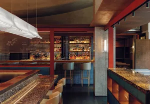 An industrial-style bar with a concrete countertop and stools underneath shelves filled with bottles of alcohol.