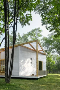 A minimalist white cabin with glass walls and wooden accents surrounded by greenery.