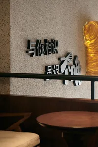 The image shows a wall art piece consisting of metallic Chinese characters displayed on a textured beige wall.