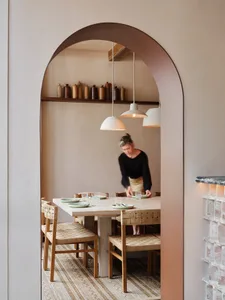 A warm-toned restaurant interior with a wooden dining table, woven chairs, and pendant lights hanging above.