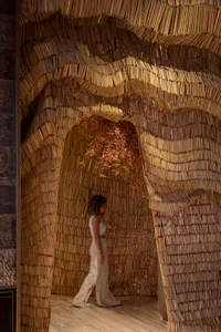 A person walking through a wave-like architectural installation made of wooden slats with dried leaves hanging from it.