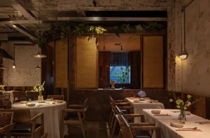 Industrially styled restaurant interior with wooden tables and chairs, white tablecloths, and green plants.