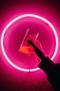 Hand holding a glass filled with a pink liquid, set against a vibrant pink neon circle background.