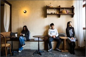 Three people sitting in a cozy cafe with wooden tables, fabric cushions, concrete walls, and metal furniture.