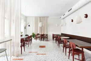 A minimalist restaurant interior with terrazzo flooring, wooden tables, and red chairs.