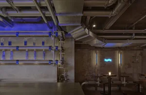 An industrial-style restaurant interior with metal pipes, concrete walls, and blue lighting.