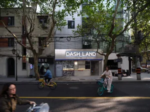A city street with people riding bicycles, a cafe with a sign that says 'DASH LAND', and trees lining the sidewalk.