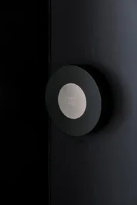 A minimalist, black, round, metallic door handle against a black, textured wall background.