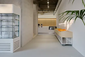 Minimalist cafe interior with white concrete walls and floors, featuring a transparent beverage display case and a wooden cou