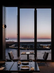 A modern restaurant interior with a sea view, featuring wooden tables and chairs, plates, and glasses set on a table.