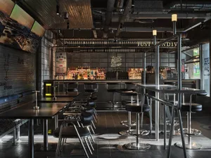 Industrial-style restaurant bar with metallic tables, stools, and a well-stocked bar area.