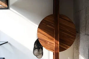A round wooden table with a mesh bag hanging from it, under a window with sunlight casting a shadow on the wall.