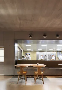 Industrial-style restaurant kitchen featuring concrete floors and walls, with chefs preparing food behind a transparent barri
